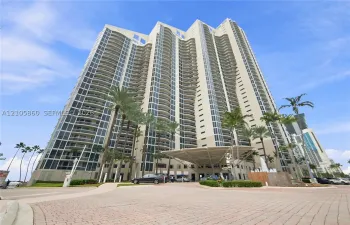 High-rise residential tower featuring a concrete facade, floor-to-ceiling windows, private balconies, a porte-cochère entry, and landscaped grounds with palm trees