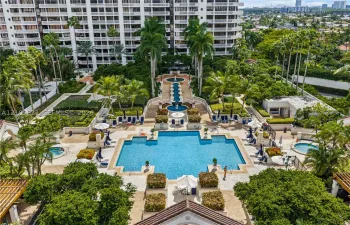 Resort-style pool area featuring a multi-tiered fountain, stone-paved sun deck, mature palm trees, and two separate hot tubs