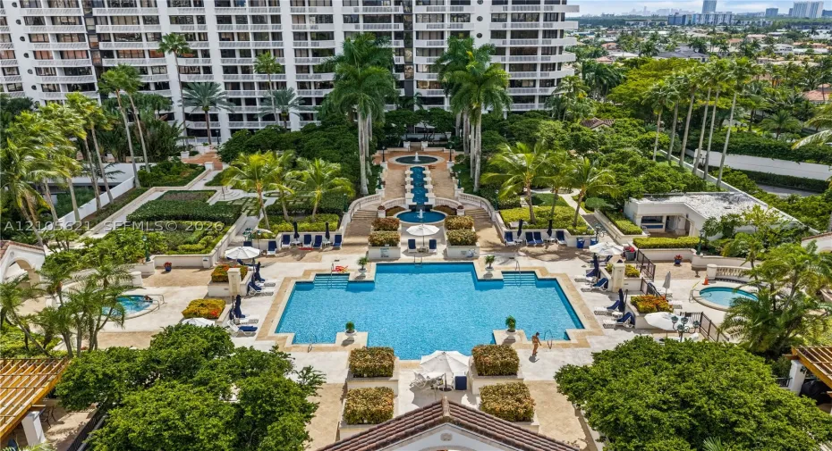 Resort-style pool area featuring a multi-tiered fountain, stone-paved sun deck, mature palm trees, and two separate hot tubs
