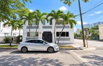 Two-story exterior facade featuring white stucco finish, recessed grey paneling, metal-grille front entryway, and integrated landscaping with mature palm trees