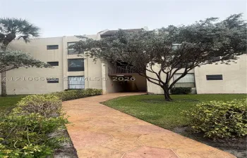Textured paver walkway leading to a multi-story building entrance with a covered balcony and surrounding mature trees and shrubs