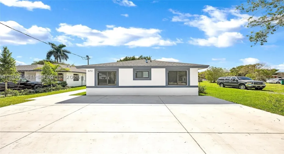 Single-story exterior featuring white stucco finish, gray trim, asphalt shingle roofing, and an expansive concrete driveway