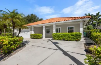 Concrete driveway leading to a residence with a terracotta tile roof, white exterior columns, and landscaping featuring palm trees and manicured hedges