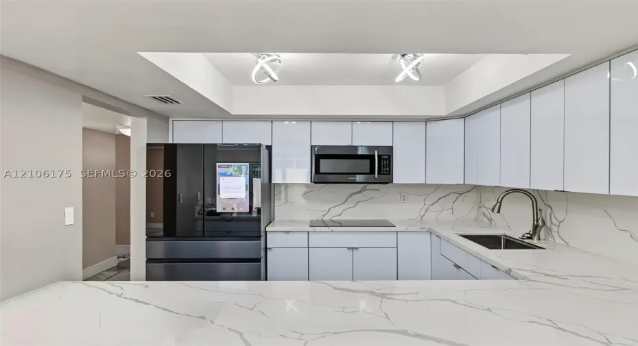 Kitchen featuring high-gloss white cabinetry, stone-patterned countertops, a matching full-height backsplash, a stainless steel undermount sink with a gooseneck faucet, and recessed tray ceiling lighting