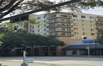 Multi-story residential building featuring a beige stucco facade, stacked corner balconies with metal railings, and ground-level commercial storefronts