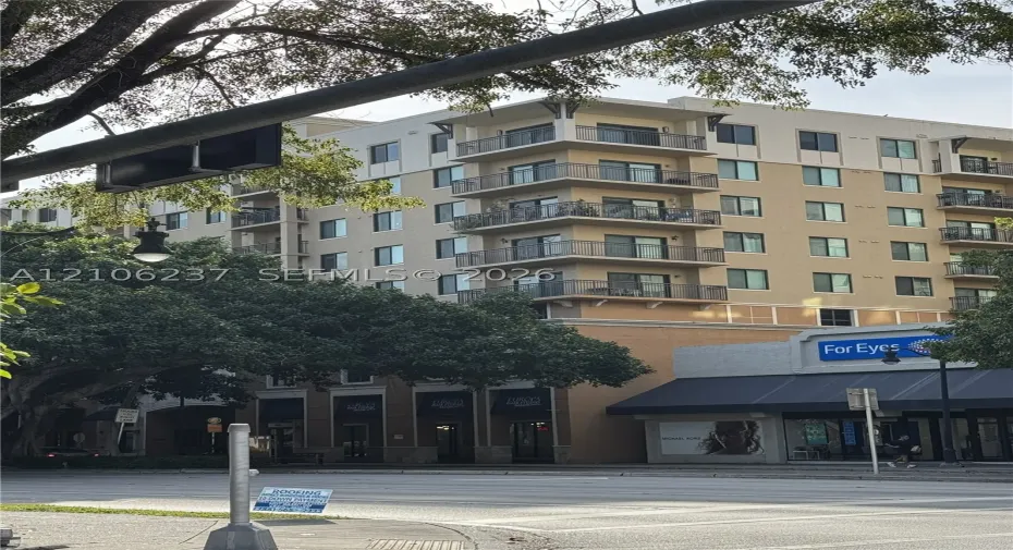 Multi-story residential building featuring a beige stucco facade, stacked corner balconies with metal railings, and ground-level commercial storefronts