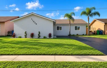 Stucco exterior with a pitched shingle roof, complemented by a paved driveway, mature palm trees, and manicured landscaping