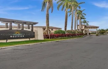 Gated community entrance featuring a modern marquee sign, architectural pergola, stone-clad gatehouse, and rows of mature palm trees