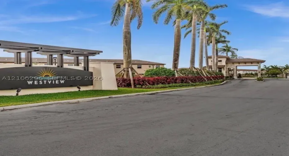 Gated community entrance featuring a modern marquee sign, architectural pergola, stone-clad gatehouse, and rows of mature palm trees