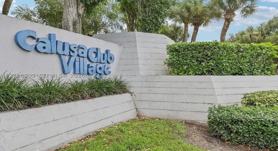 Monument signage for Calusa Club Village featuring a tiered concrete structure, integrated landscaping, and mature palms