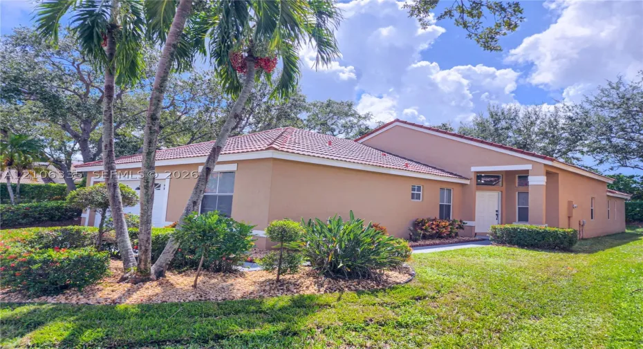 Stucco exterior featuring a hipped tile roof, integrated garage, covered entryway, and mature tropical landscaping