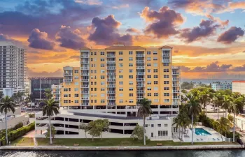 Multi-story building featuring private balconies, integrated parking structure, waterfront positioning, surrounding mature palm trees, and an outdoor swimming pool
