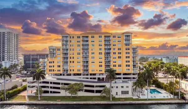 Multi-story building featuring private balconies, integrated parking structure, waterfront positioning, surrounding mature palm trees, and an outdoor swimming pool