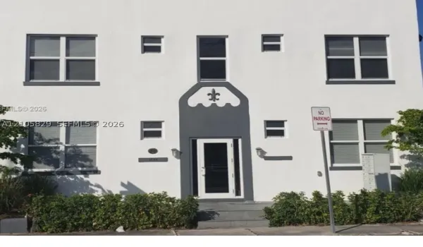 White stucco exterior featuring a decorative fleur-de-lis motif above the entryway, multi-pane windows with dark framing, and wall-mounted exterior lighting
