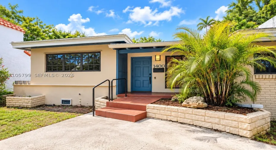 Single-story exterior featuring a light-toned stucco facade, tiered stone garden retaining walls, a covered entryway with tile steps, and a blue paneled front door
