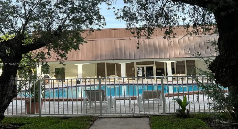 Swimming pool with brick coping, surrounded by a paved deck and enclosed by a metal perimeter fence, featuring a covered patio with structural columns