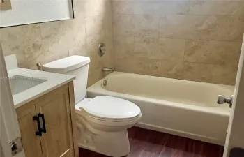 Bathroom featuring wood-finish vanity cabinetry, solid-surface countertop with integrated sink, wood-finish flooring, and a bathtub surround finished in rectangular stone-style tile