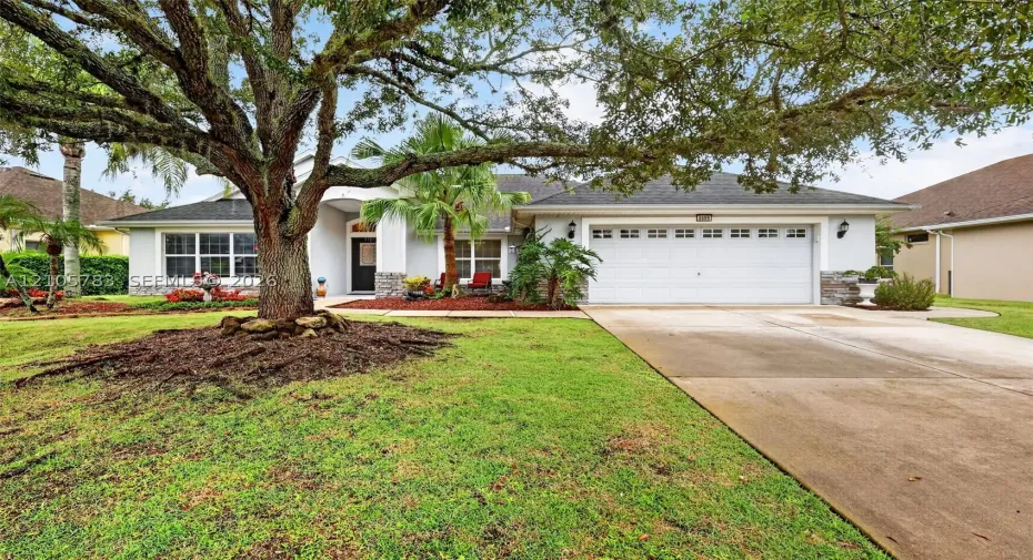 Single-story exterior featuring a two-car garage, stone veneer accents, multi-pane windows, and a mature oak tree in the front lawn