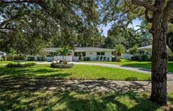 Single-story exterior featuring light-toned stucco siding, an expansive front lawn, mature canopy trees, and a concrete driveway