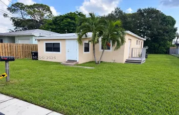 Stucco exterior residence featuring a flat roofline, white-framed windows, a wood-constructed side deck, and mature palm trees situated on an expansive grass lot