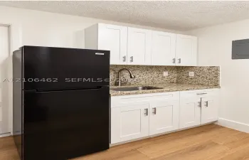White shaker-style cabinetry with satin nickel hardware, stone-composite countertops, stainless steel undermount sink with gooseneck faucet, black top-freezer refrigerator, and wood-finish flooring