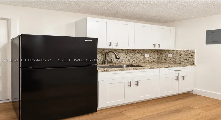 White shaker-style cabinetry with satin nickel hardware, stone-composite countertops, stainless steel undermount sink with gooseneck faucet, black top-freezer refrigerator, and wood-finish flooring