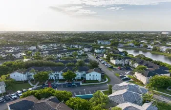 Aerial perspective of a multi-unit residential community featuring shingle-style roofing, paved parking areas, sidewalk pathways, and a water feature
