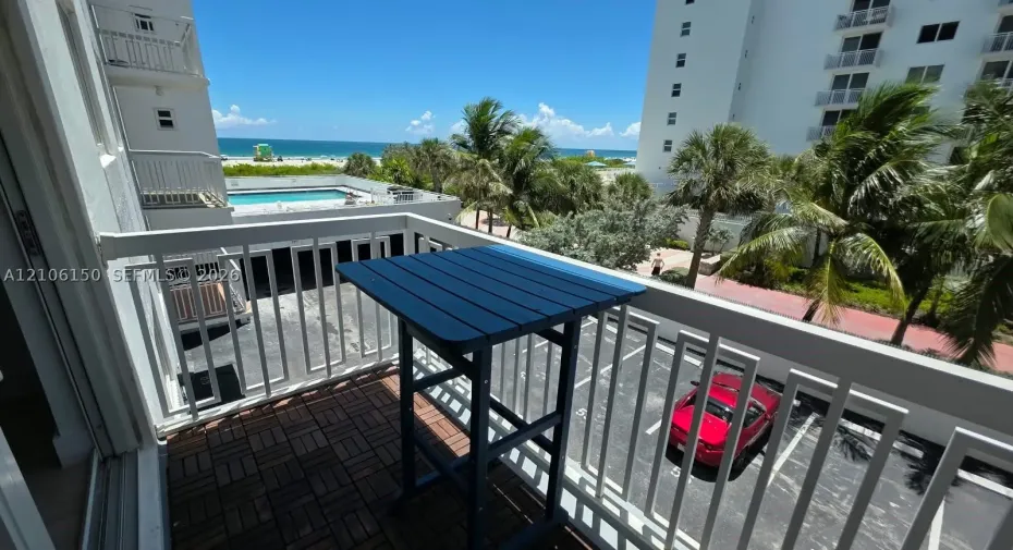 Private balcony featuring white metal railings and interlocking deck tiles, overlooking the coastline and community pool