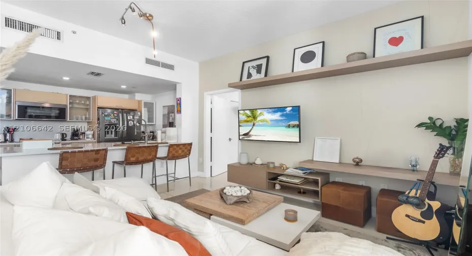 Living area featuring custom floating shelves, built-in cabinetry, and neutral-toned wall treatments, paired with an open kitchen containing integrated appliances, light-wood cabinetry, and stone-surfaced breakfast bar