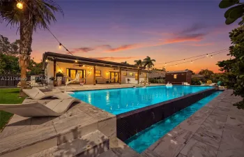 Expansive outdoor area featuring a large swimming pool with an integrated waterfall, a spacious patio with multiple access points to the residence, and mature tropical landscaping
