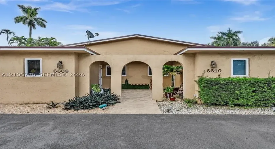 Single-story duplex exterior featuring a central covered entryway with triple arches, stone-paved walkway, stucco finish, and wall-mounted exterior light fixtures