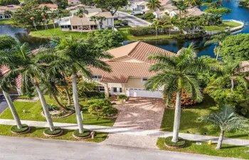 Two-story residence featuring a tile roof, brick-paver driveway, multi-car garage, mature palm landscaping, and waterfront positioning