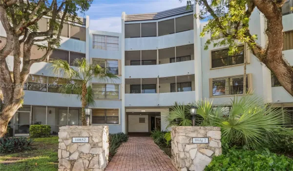 Multi-story building exterior featuring a brick paver walkway, stacked stone pillars, screened-in balconies, and mature landscaping