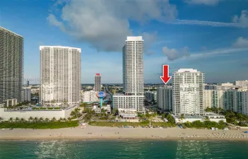 Aerial perspective of a high-rise residential building situated along a coastal shoreline, featuring direct beach access and proximity to regional urban centers