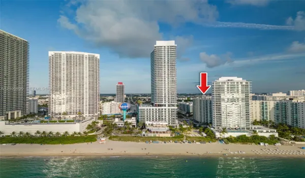 Aerial perspective of a high-rise residential building situated along a coastal shoreline, featuring direct beach access and proximity to regional urban centers