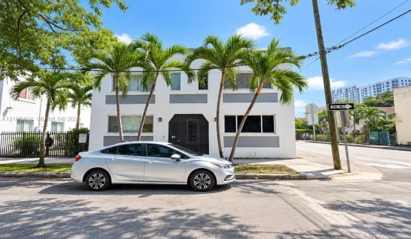 Two-story exterior facade featuring white stucco finish, recessed grey paneling, metal-grille front entryway, and integrated landscaping with mature palm trees