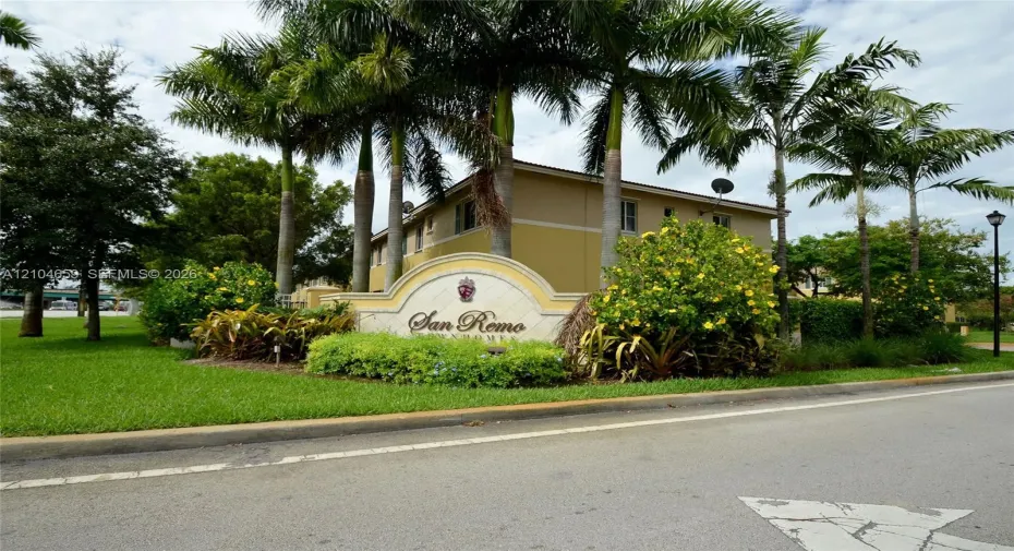 Community entrance featuring a monument sign, manicured lawn, mature palm trees, and native flowering shrubs