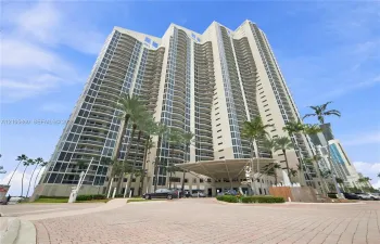 High-rise residential tower featuring a concrete facade, floor-to-ceiling windows, private balconies, a porte-cochère entry, and landscaped grounds with palm trees