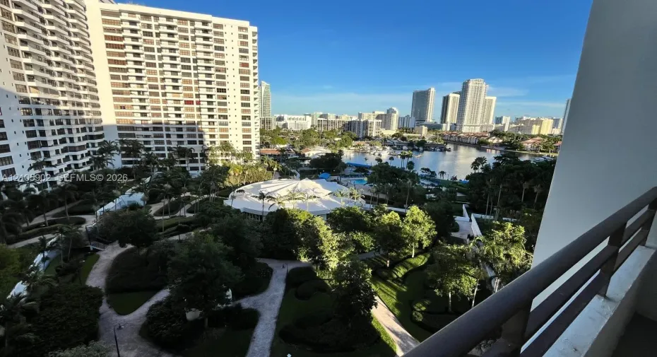 Balcony featuring horizontal metal railings, overlooking a landscaped garden, paved walkways, and water-front cityscape