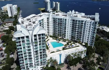 High-rise waterfront property featuring a large swimming pool, expansive paved deck, glass-railed private balconies, and mature palm landscaping