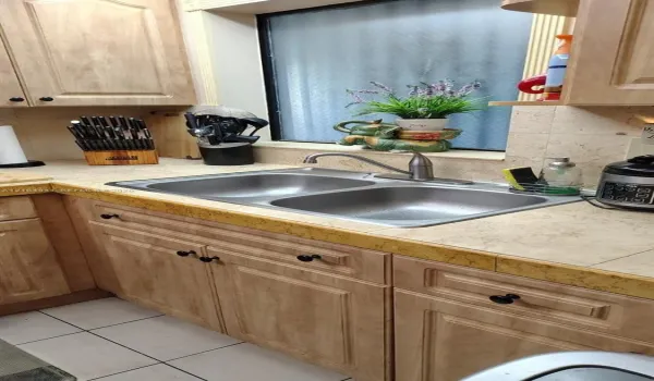 Light wood-finish cabinetry featuring raised-panel doors, neutral tiled countertops with decorative trim, and a double-basin stainless steel sink with a high-arch faucet