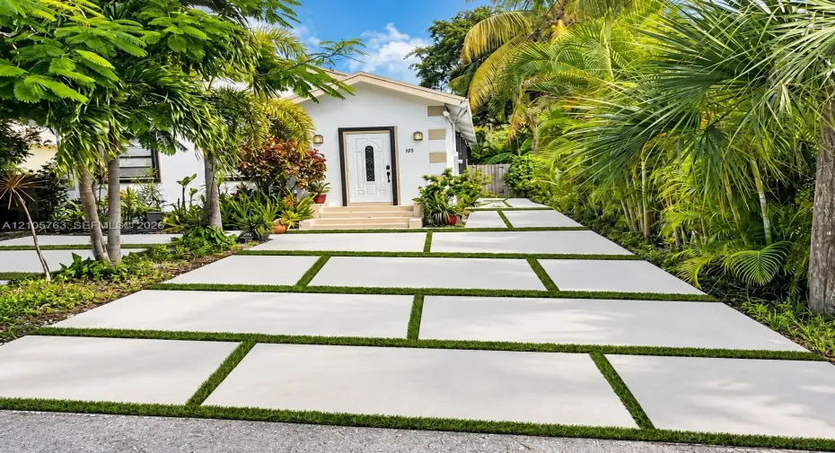 Exterior facade featuring white stucco walls, decorative quoins, a white paneled front door with glass inlay, and a concrete paver driveway with integrated turf strips
