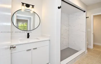 White vanity with stone-composite countertop, undermount sink, matte black faucet, round mirror with black frame, and walk-in shower featuring large-format patterned wall tile and dark hexagonal floor tile