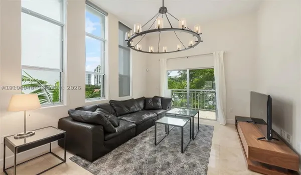 High-ceilinged living space featuring floor-to-ceiling windows, a large metal circular chandelier, stone flooring, and a glass-paneled balcony door