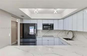 Kitchen featuring high-gloss white cabinetry, stone-patterned countertops, a matching full-height backsplash, a stainless steel undermount sink with a gooseneck faucet, and recessed tray ceiling lighting