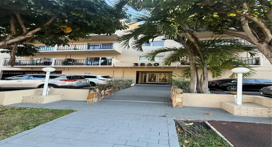 Multi-story building facade featuring private balconies, masonry retaining walls, a paved walkway, and mature palms