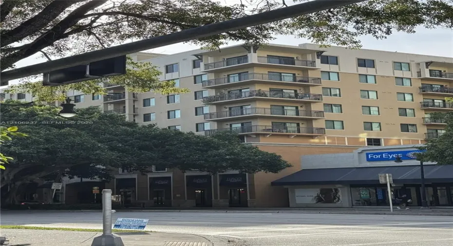 Multi-story residential building featuring a beige stucco facade, stacked corner balconies with metal railings, and ground-level commercial storefronts