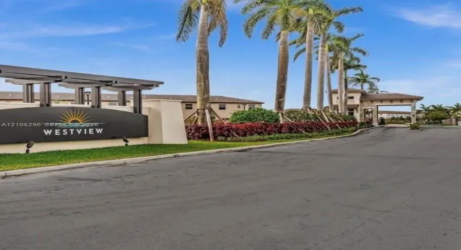 Gated community entrance featuring a modern marquee sign, architectural pergola, stone-clad gatehouse, and rows of mature palm trees