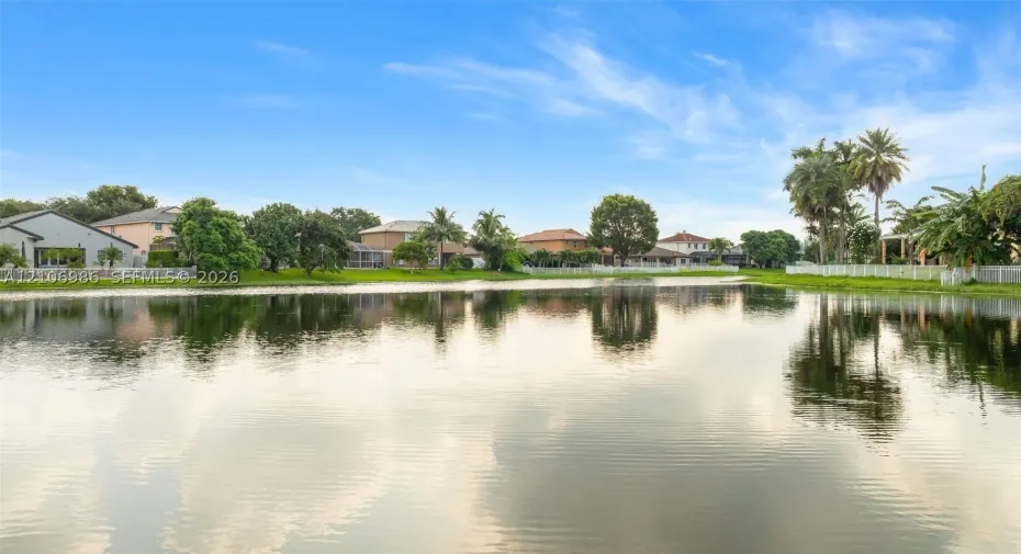 Waterfront setting featuring a calm pond, mature palm trees, perimeter white fencing, and expansive green space