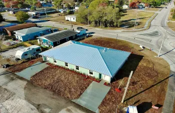 Single-story structure featuring a standing-seam metal roof, stucco exterior, paved driveway, and established landscape beds